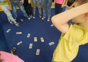 Dzieci stoją, a na dywanie znajdują się banknoty (falsyfikaty) i monety.