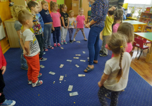 Dzieci stoją, a na dywanie znajdują się banknoty (falsyfikaty) i monety.