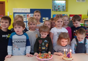 Dzieci stoją przy stole. Jedna dziewczynka siedzi przed tortem na którym płoną świeczki.