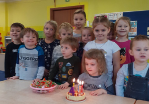 Dzieci stoją przy stole. Jedna dziewczynka siedzi przed tortem na którym płoną świeczki
