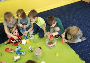 5 dzieci siedzą na dywanie i wsypują do puszek różne materiały.