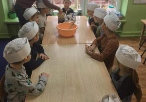 Dzieci siedzą przy stole w kucharskich czapkach.