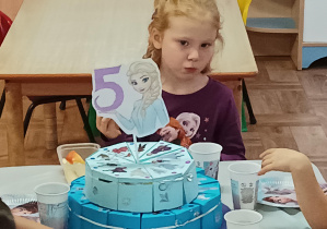 dziewczynka siedzi przy stoliku, na stole stoi urodzinowy tort, widać napis 5 lat