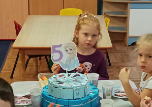 dziewczynka siedzi przy stoliku, na stole stoi urodzinowy tort, widać napis 5 lat