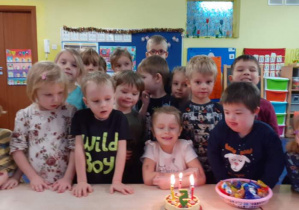 Dzieci stoją wokół stołu, a dziewczynka siedzi przed tortem ze świeczkami.