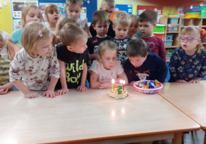 Dzieci stoją wokół stołu, a dziewczynka siedzi przed tortem ze świeczkami.