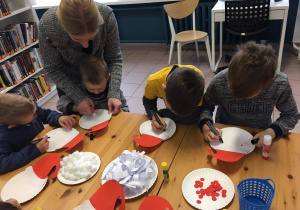 Dzieci siedzą przy stole i wykonują portret Mikołaja z papierowego talerzyka.