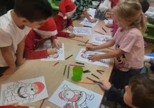 Dzieci siedzą przy stole i kolorują Św. Mikołaja.