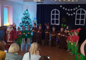 Występ dzieci przed rodzicami z okazji świąt Bożego Narodzenia. Na sali dzieci przebrane za renifery, elfy oraz Mikołaja śpiewają piosenki, recytują wiersze, grają na instrumentach, tańczą.