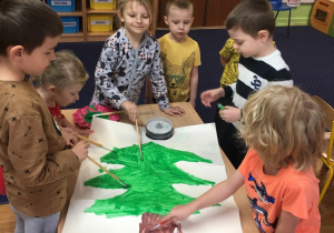 Dzieci stoją wokół stołu i malują choinkę farbami.