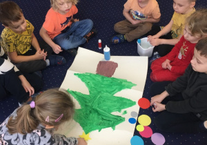 Dzieci siedzą na dywanie przed nimi leży pokolorowana choinka. Dziewczynka przykleja gwiazdę na czubku choinki.