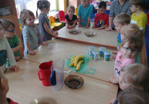 Dzieci stoją przy stołach, na których znajdują się banany, jogurty, blendery.