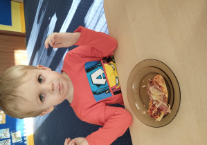 chłopiec siedzi przy stoliku, przed nim na talerzu widoczny jest trójkątny kawałek pizzy