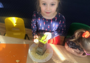 Dziewczynka przy stole ściera żółty ser na tarce.