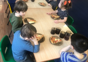 Dzieci siedzą przy stole i zjadają własnoręcznie wykonaną pizzę.