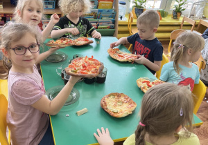 Dzieci siedzą przy stole i zjadają samodzielnie skomponowaną pizzę.