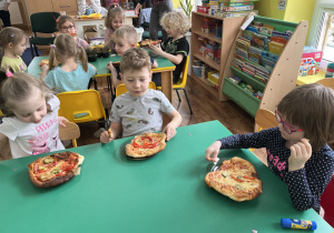 Dzieci siedzą przy stole i zjadają samodzielnie skomponowaną pizzę.