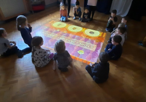 Dzieci siedzą na sali gimnastycznej i obserwują podłogę interaktywną.