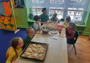 Dzieci siedzą przy stolikach, a przed nimi pizze gotowe do upieczenia.