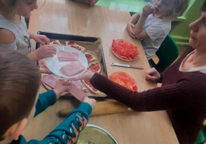 Dzieci siedzą przy stolikach i przygotowują pizzę.