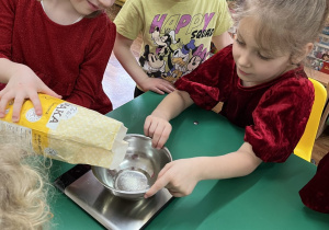 Dzieci zgromadzone przy stole. Dziewczynka odmierza odpowiednią ilość mąki.