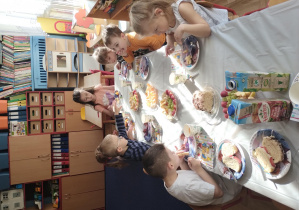 dzieci siedzą przy stolikach, na których ustawione są talerzyki ze smakołykami