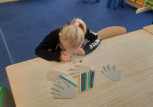 Dziewczynka siedzi przy stoliku i maluje papierowe paznokcie..