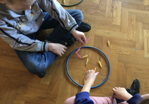 Dzieci siedzą na podłodze w parach i łowią słomką makaron.
