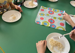 Dzieci siedzą przy stole i nawijają spaghetti na widelec.
