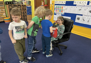 Dzieci ustawione w rzędzie podchodzą do dziewczynki siedzącej na krześle i składają jej życzenia.