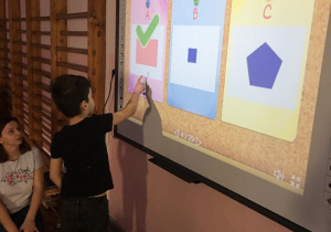 Chłopiec stoi przed tablicą multimedialną i wskazuje prostokąt.