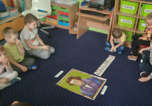 Dzieci siedzą na dywanie w kole. Na środku leży napis "kompozytor" i portret Fryderyka Chopina.