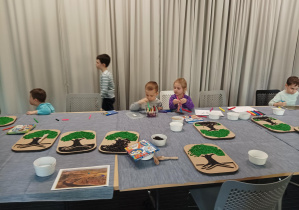 dzieci z przedszkola uczestniczą w warsztatach sensorycznych organizowanych w bibliotece, dzieci siedzą przy stolikach i wykonują prace manualne, wsypują kolorowe ziarna do szablonu drzewa