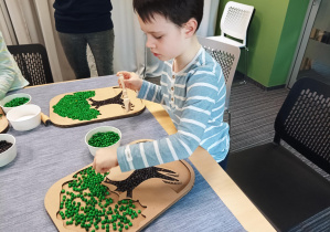 dzieci z przedszkola uczestniczą w warsztatach sensorycznych organizowanych w bibliotece, dzieci siedzą przy stolikach i wykonują prace manualne, wsypują kolorowe ziarna do szablonu drzewa