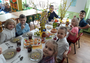 Dzieci siedzą przy stole na którym są smakołyki i biały barszcz.