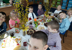 Dzieci siedzą przy stole na którym są smakołyki i biały barszcz.