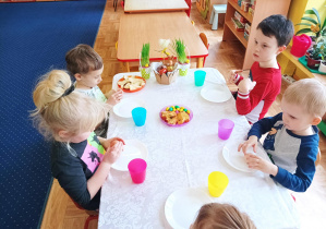 dzieci siedzą przy stoliku nakrytym białym obrusem, częstują się słodyczami