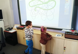 chłopiec i dziewczynka stoją przy tablicy multimedialnej w bibliotece