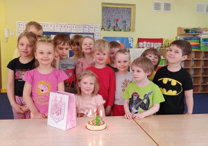 grupa dzieci stoi za stołem, przy stole siedzi dziewczynka, przed nią widać zabawkowy tort z cyfrą 5