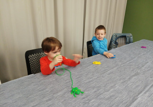 dzieci siedzą przy dużym stole, chłopiec trzyma w ręku zabawę żabkę