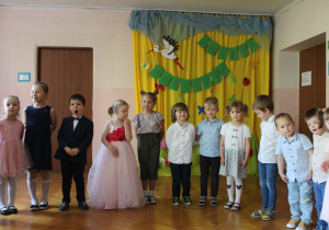grupa dzieci w odświętnych strojach prezentuje wiersze dla rodziców, stoją na sali gimnastycznej