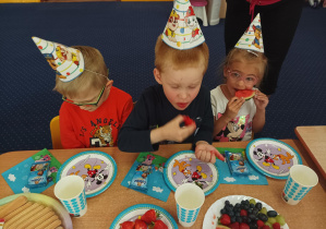 dzieci w urodzinowych czapeczkach siedzą przy stolikach, na nich stoją talerze z poczęstunkiem