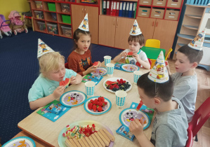 dzieci w urodzinowych czapeczkach siedzą przy stolikach, na nich stoją talerze z poczęstunkiem