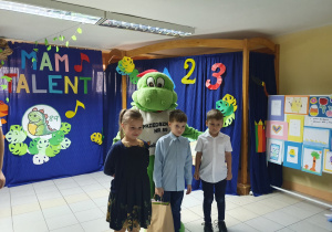 Troje dzieci wraz z maskotką dinozaura pozuje do zdjęcia.