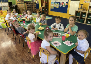 Dzieci siedzą przy stole na którym stoi słodki poczęstunek.