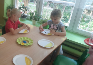 Dzieci siedzą przy stole i malują farbami talerzyki.