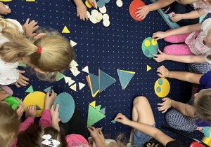 Dzieci siedzą na dywanie i układają kompozycje z figur geometrycznych.