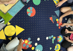 Dzieci siedzą na dywanie i układają kompozycje z figur geometrycznych.