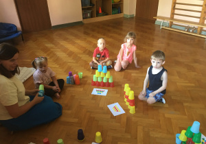 Dzieci siedzą na podłodze i bawią się kolorowymi kubeczkami.