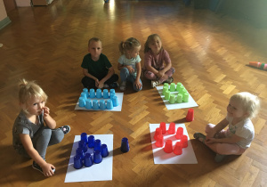 Dzieci siedzą na podłodze i układają kubeczki na figurach geometrycznych.